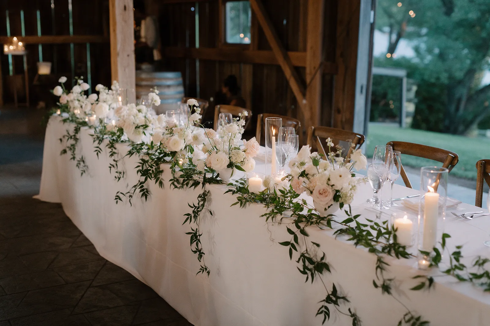 A long wedding table in a barn dressed with low arrangements of white and blush flowers, trailing greenery and candles in soft evening light