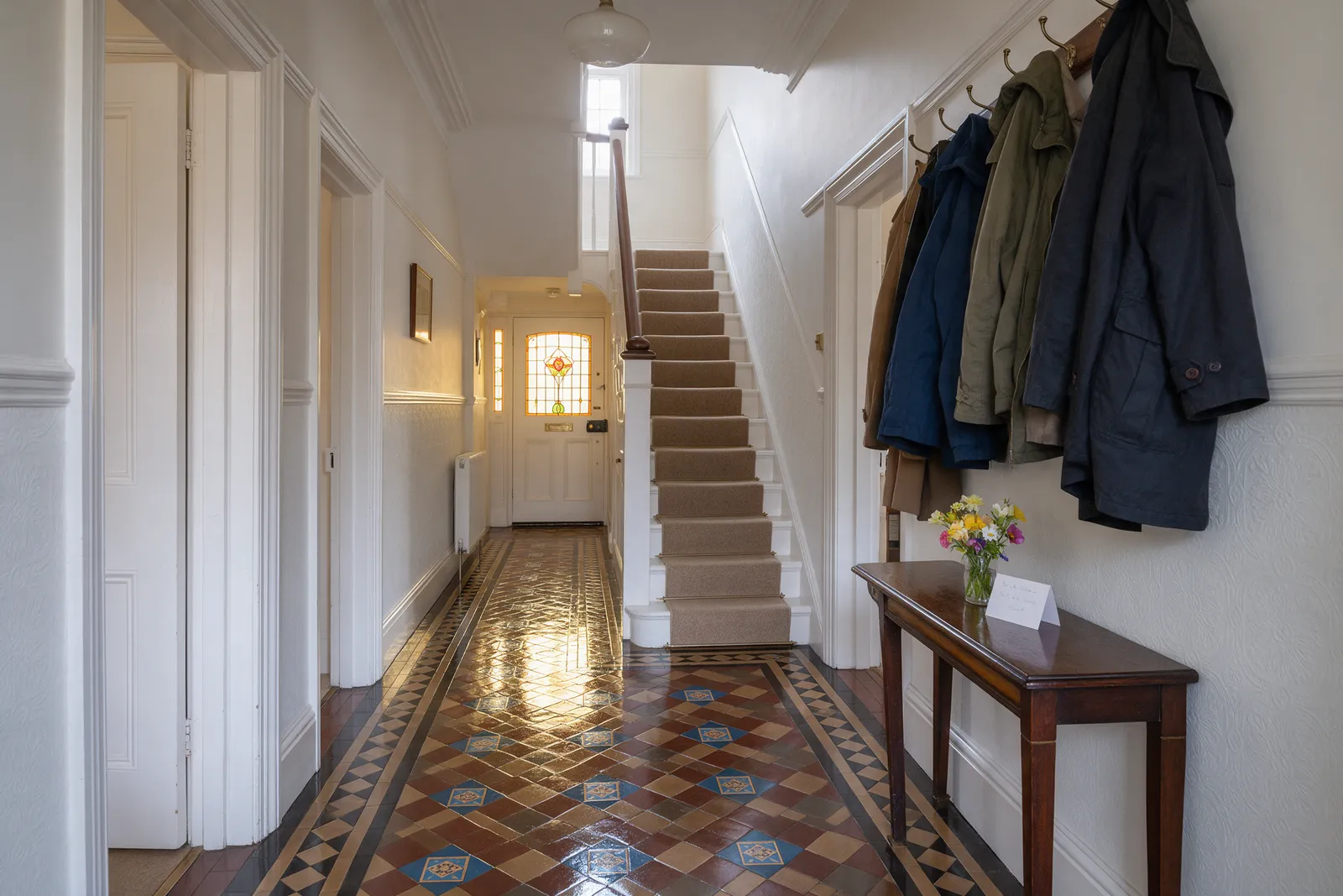 A tidy hallway in a 1930s house after cleaning with a gleaming tiled floor, a console table with fresh flowers and a card, and a clean stair runner