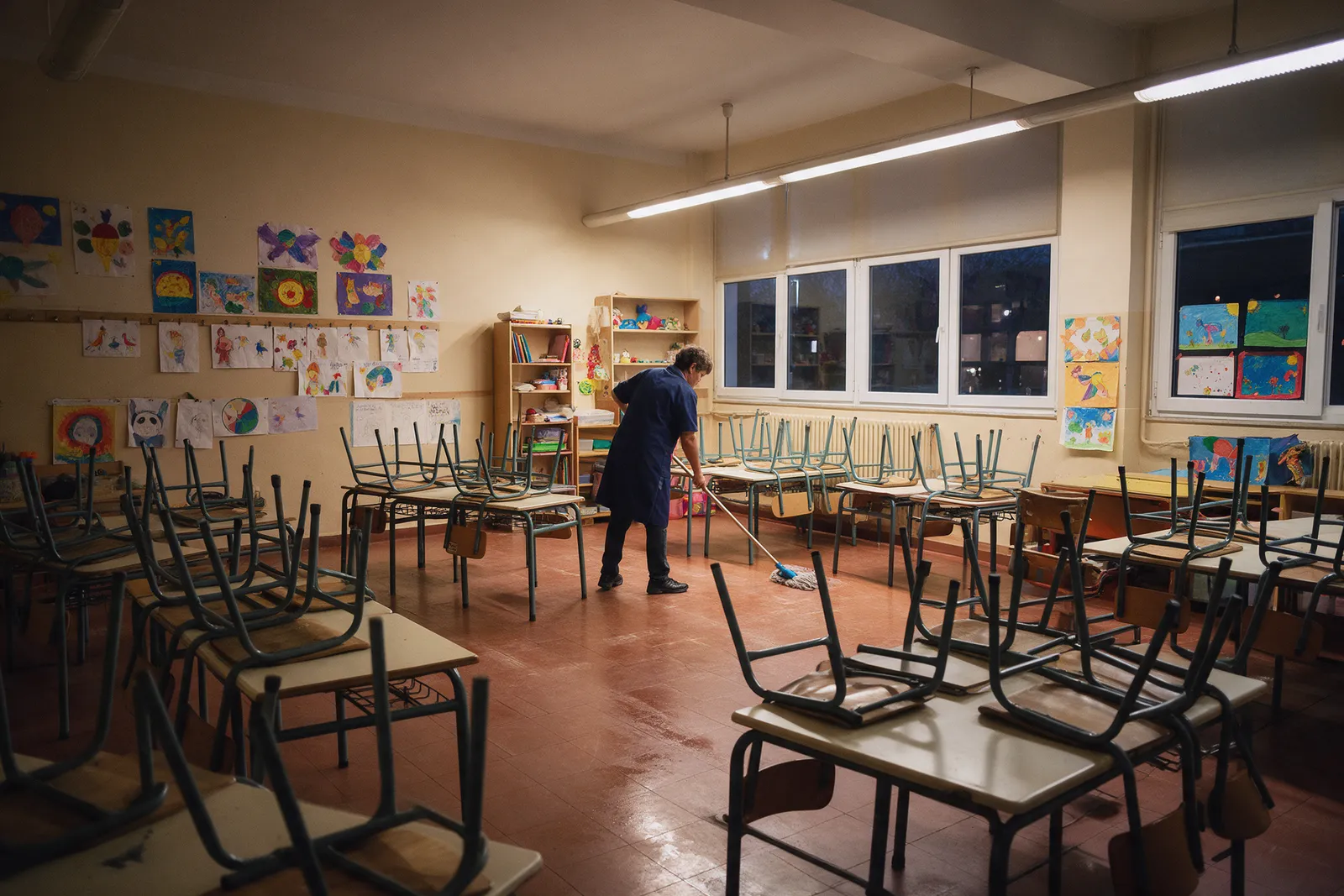 A primary school classroom after cleaning in the evening, small chairs stacked on tables and a cleaner in a navy tunic mopping the floor beneath children's artwork