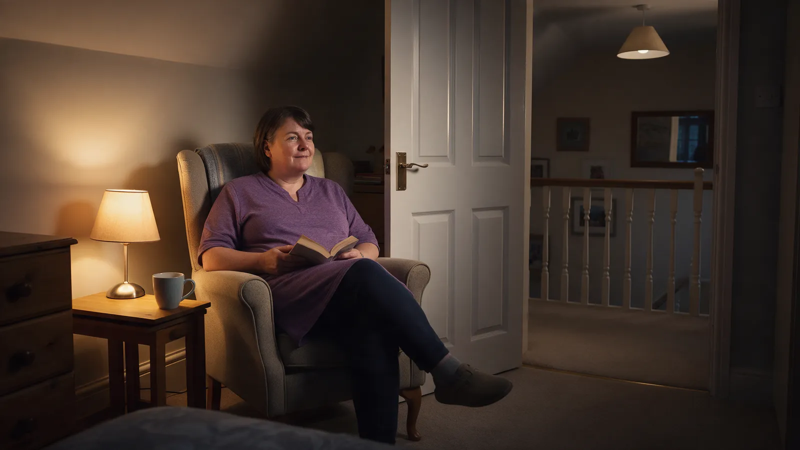 A carer sitting in a lamp-lit armchair on a quiet landing at night with a mug and a book, a bedroom door ajar beside her