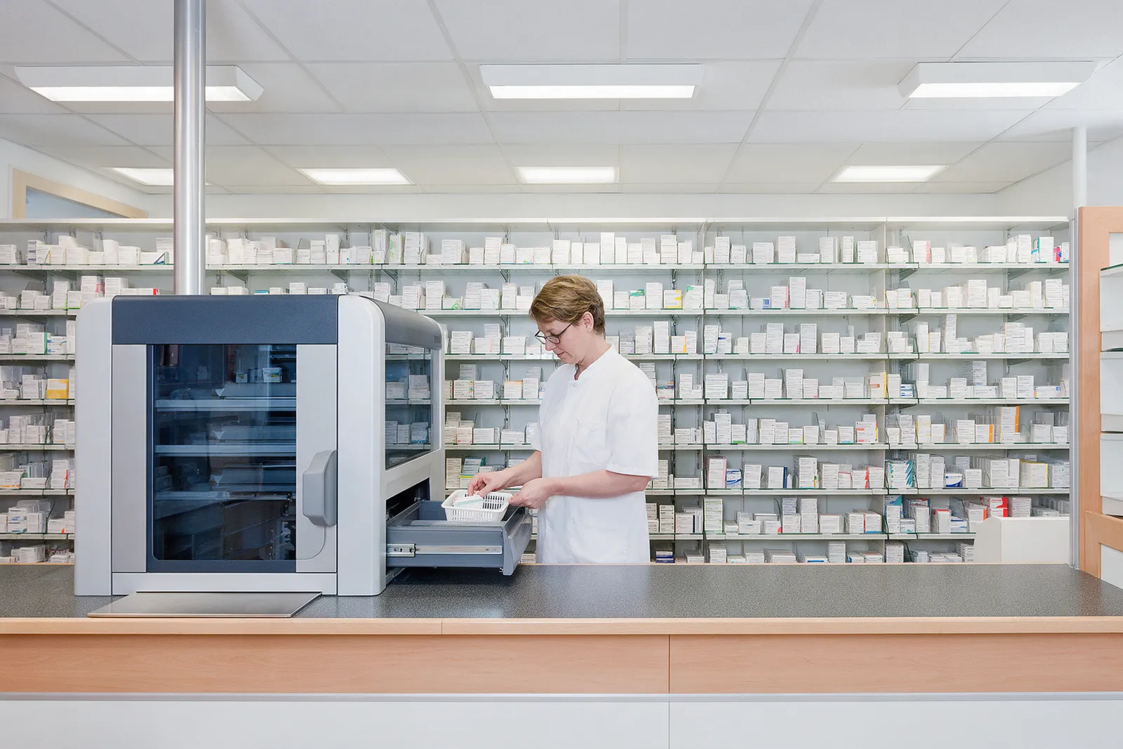 A pharmacy dispensary from behind the counter: shelves of white medicine boxes, a pharmacist checking a prescription basket, a dispensing robot drawer open