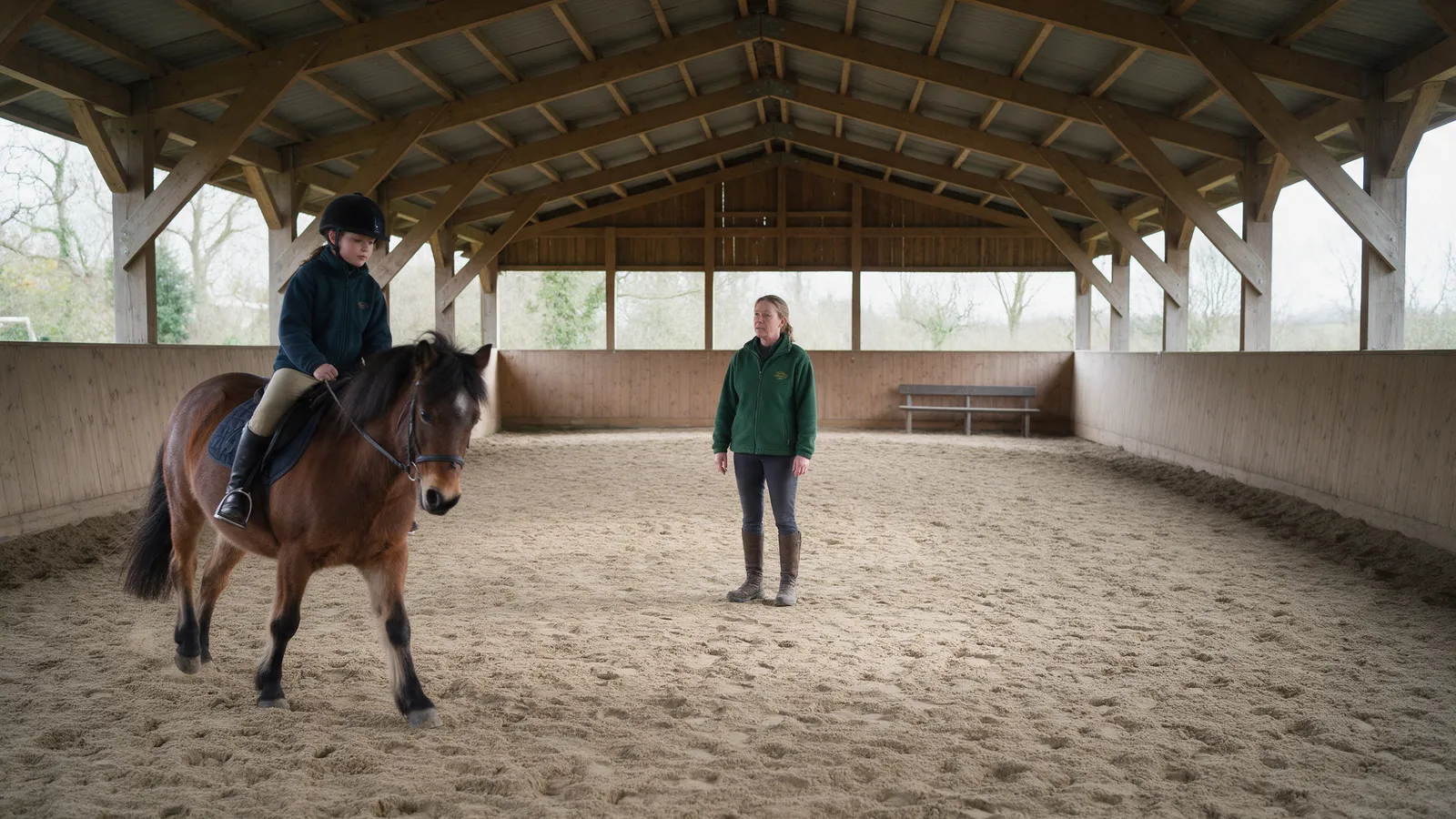 Inside a timber-framed covered riding school, a young rider on a bay pony at walk and an instructor in a green fleece in the middle