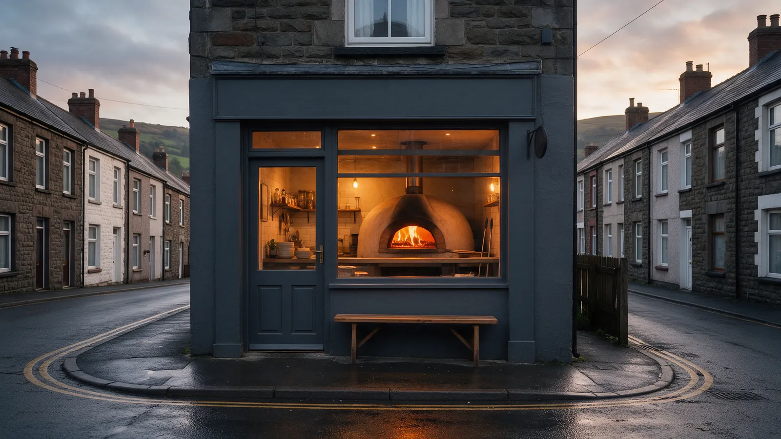 A charcoal-painted pizza shop front on a Welsh town high street at dusk, a warm orange glow from the oven through the window, a bench outside