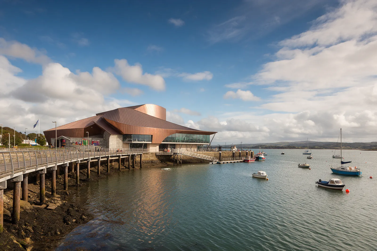 A waterfront bay with a modern copper-roofed arts centre, a boardwalk and moored boats under a bright sky