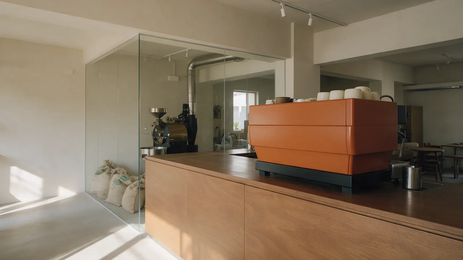 The roastery café: oat walls, an espresso-brown wooden counter, a burnt-orange espresso machine and a drum roaster behind a glass partition