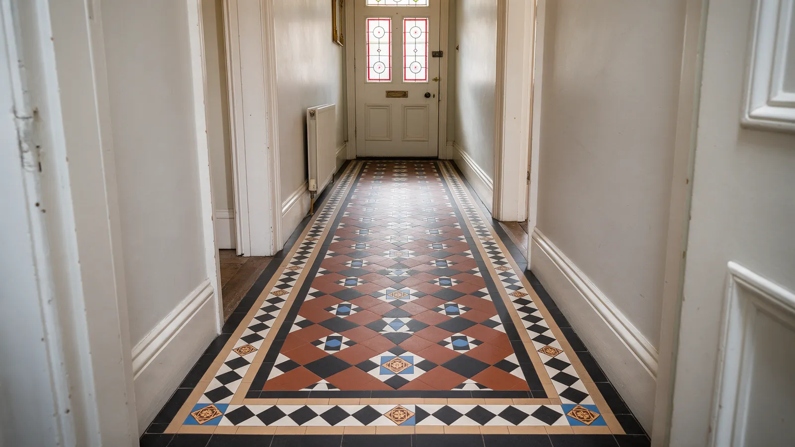 A restored Victorian hallway floor with geometric terracotta, black and blue encaustic tiles in a border pattern, leading to a stained glass front door