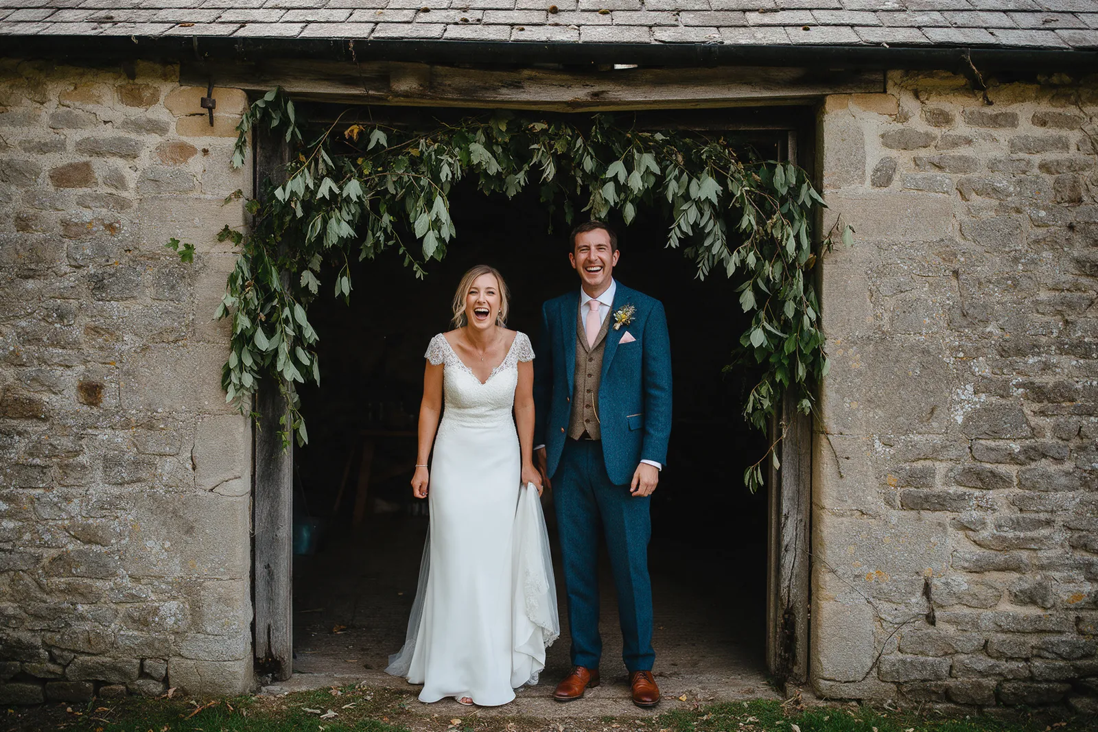 A bride and groom laughing in the doorway of an old stone barn hung with foliage in soft natural light