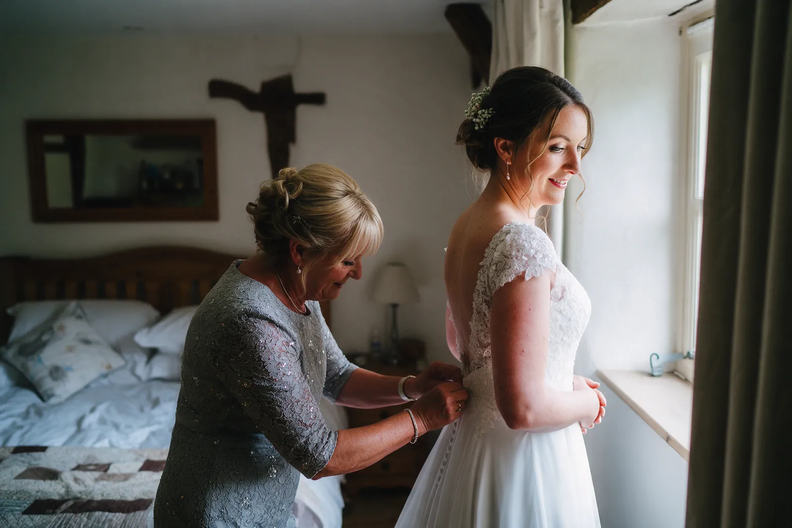 A bride having her dress buttoned by her mother by a window