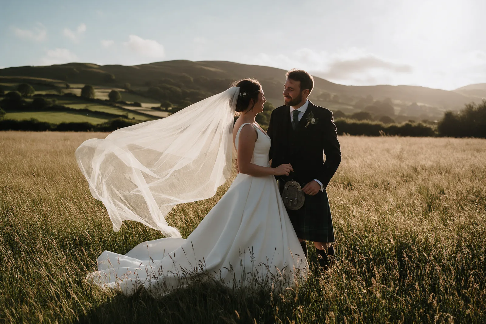 A couple standing in a long-grass meadow at golden hour with rolling Welsh hills behind, the bride's veil lifting in the wind
