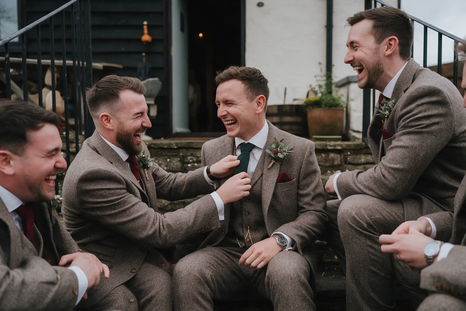 A groom and three groomsmen laughing on a farmhouse step in the morning, one adjusting another's tie badly