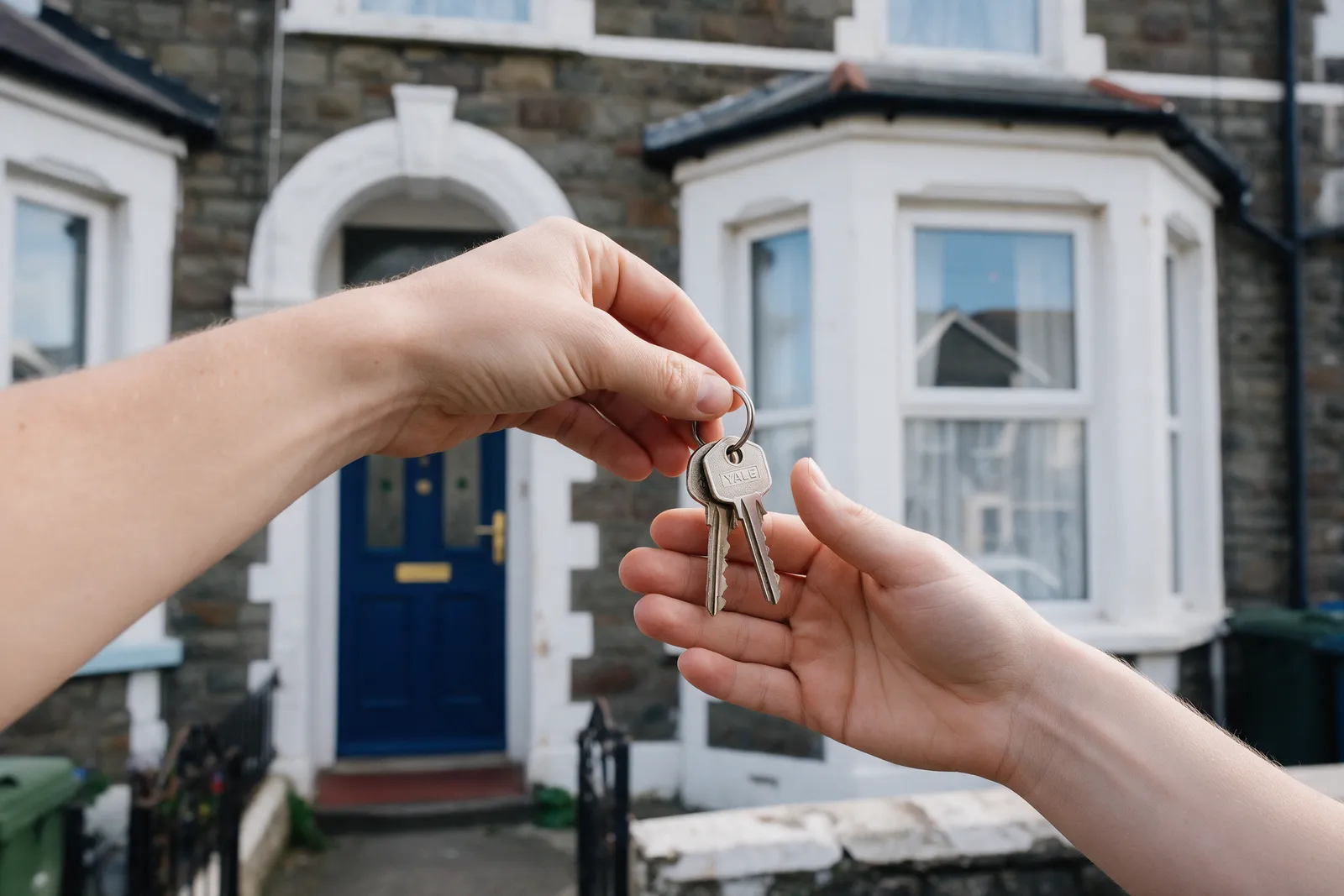 A set of house keys being handed from one hand to another at the front door of a Victorian terraced house with a bay window
