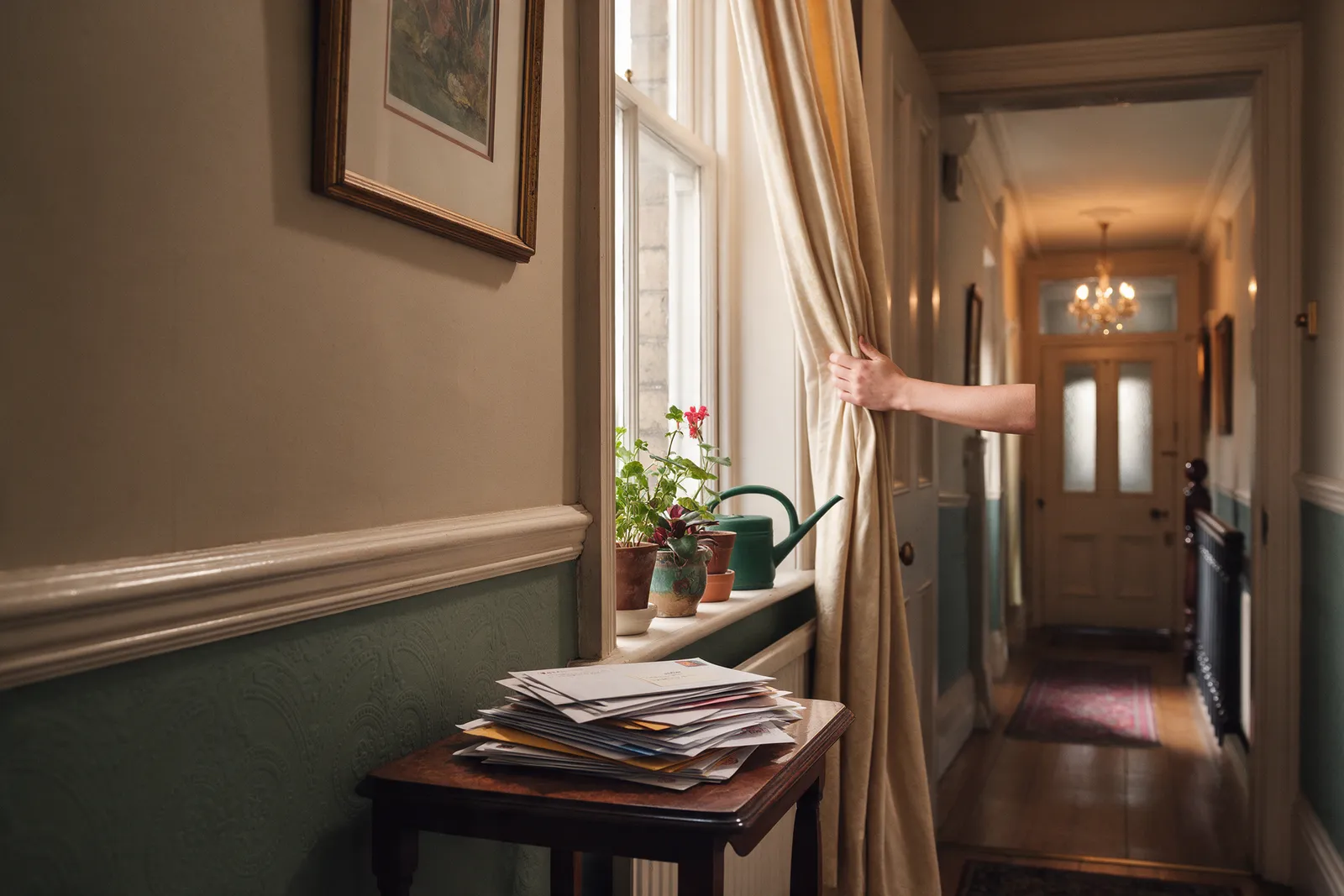 A Victorian hallway with post gathered on a side table, a watering can beside house plants, a hand drawing the curtains