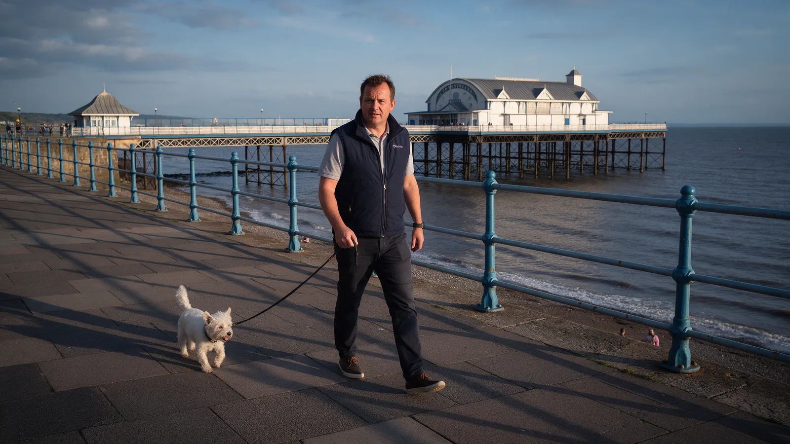 A man in a navy gilet walking a small white dog along the Penarth seafront esplanade with the pier and pavilion behind