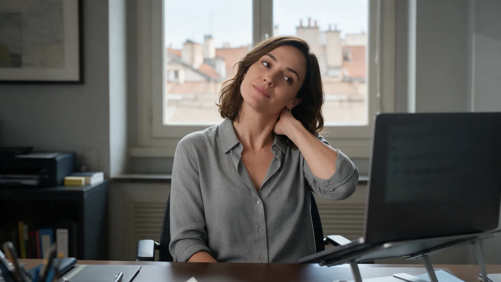 A woman in an office stretching her neck at a desk with a laptop on a stand beside a window overlooking city rooftops