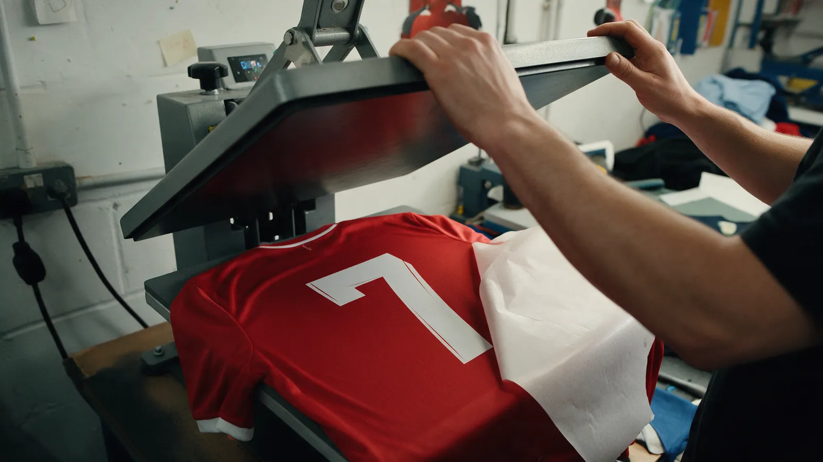 A print room worker lifting the arm of a heat press to reveal a white number freshly pressed onto the back of a red football shirt