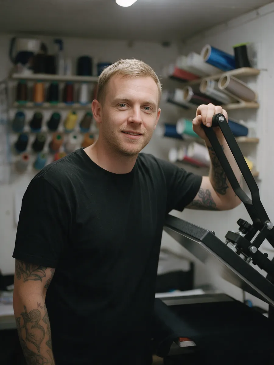 Rhys Thomas, a man in his early thirties with short fair hair and tattooed forearms, in a black T-shirt, lifting the arm of a heat press in a print room