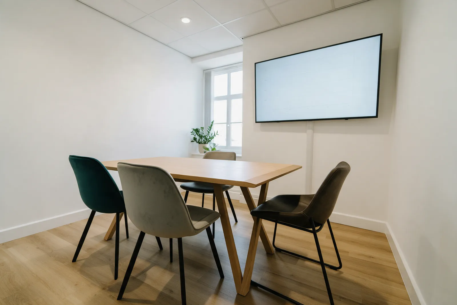A bright meeting room in a small accountancy office with a light wood table, four chairs, a large blank monitor on the wall, a plant and a window
