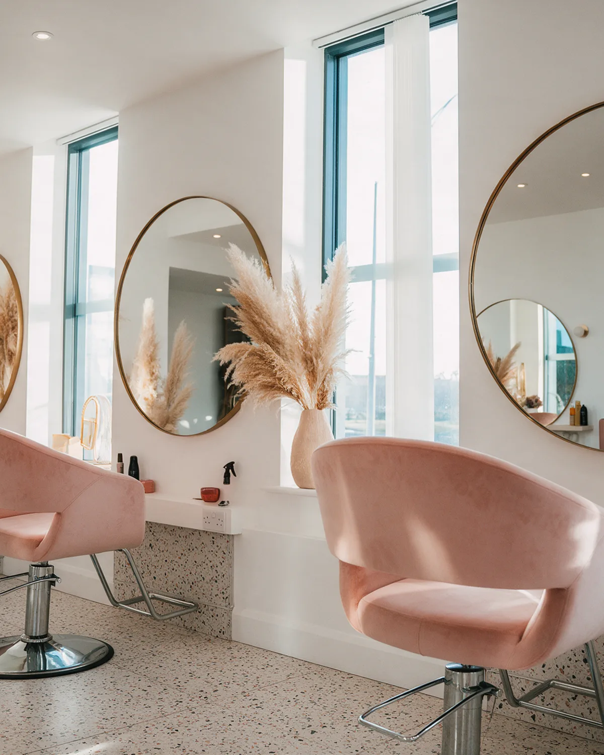 The salon: white walls, blush velvet styling chairs, round brass mirrors and afternoon light through tall windows
