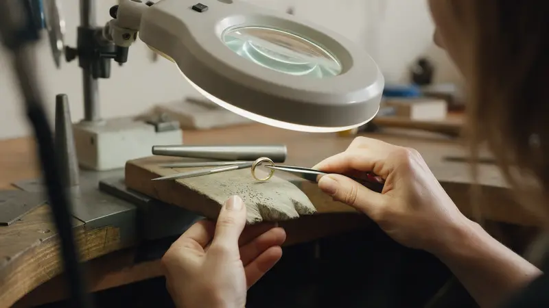 A goldsmith's hands holding a gold ring in tweezers under a bench magnifier, fine files and a ring mandrel on a worn wooden bench pin, warm lamp light