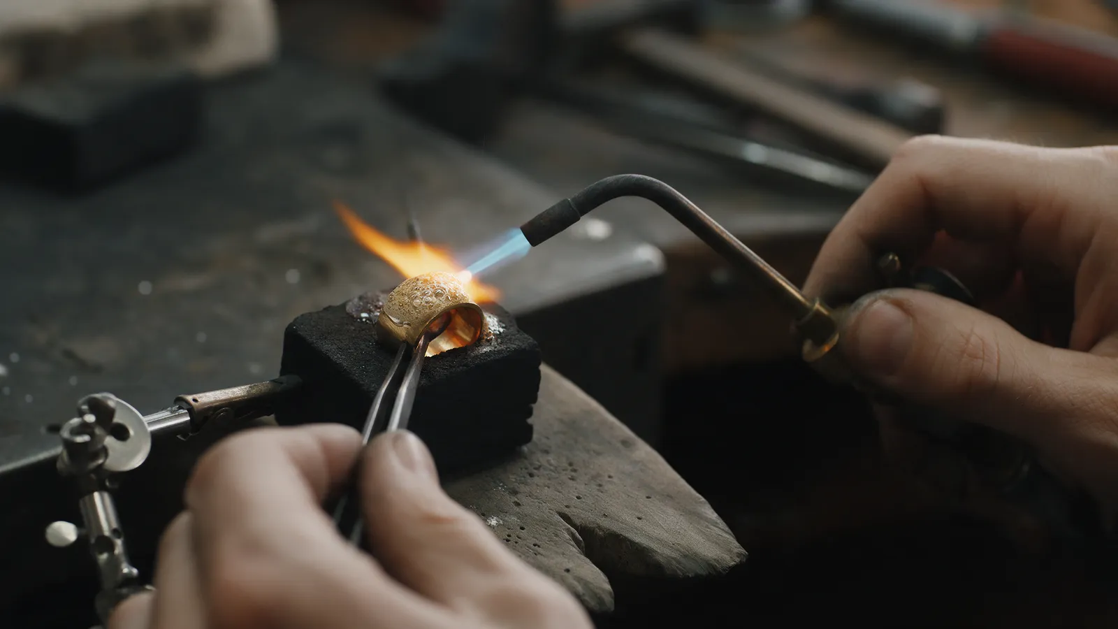 A jeweller soldering a gold ring shank with a small torch flame on a charcoal block, flux bubbling, tweezers holding the ring