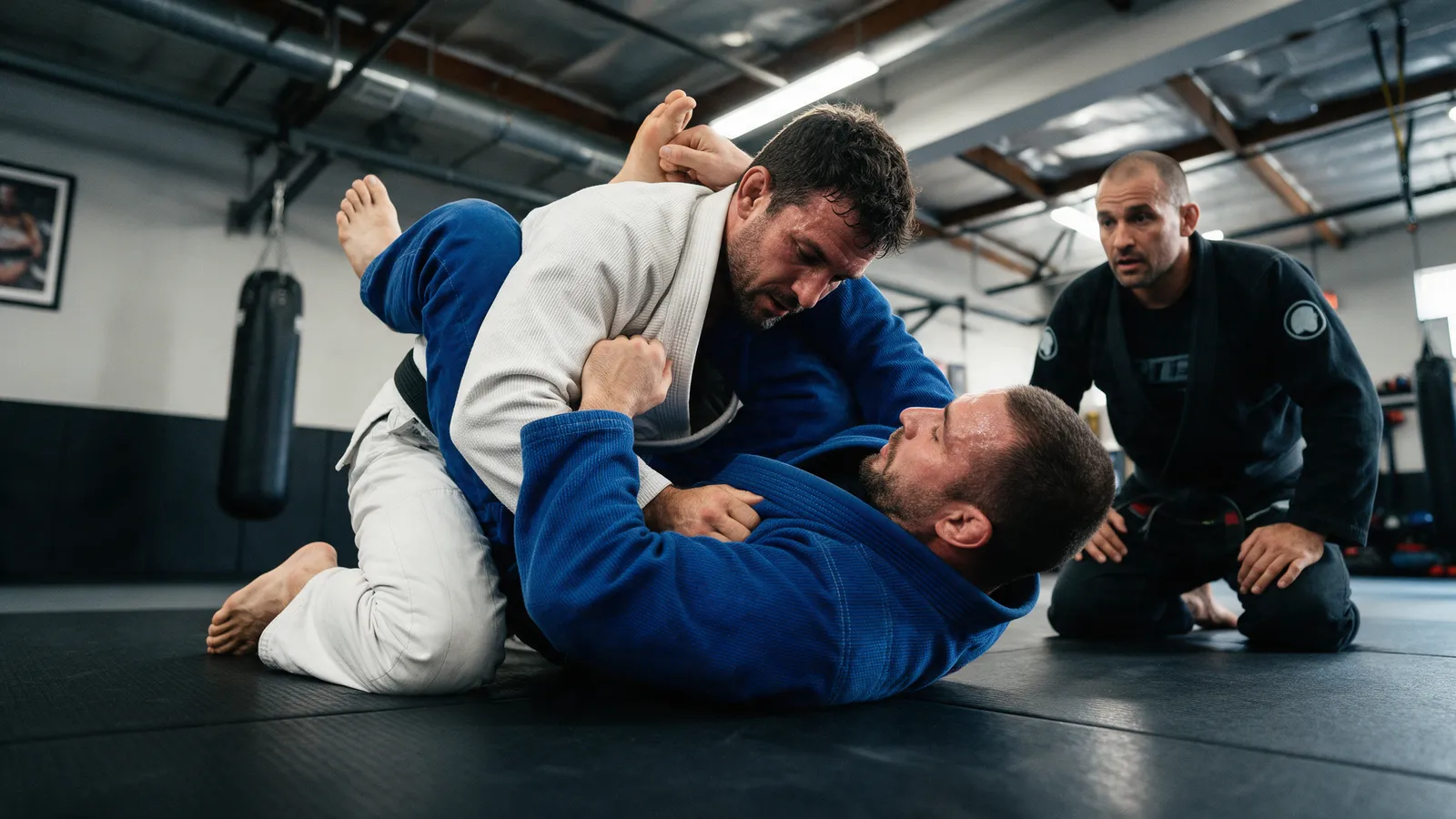 Two adults in white and blue gis grappling on black mats, one in guard, a coach kneeling nearby watching closely