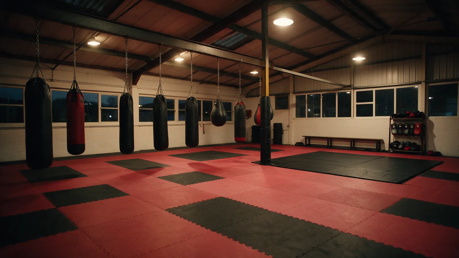 The inside of an empty martial arts gym in an industrial unit with red and black mats, heavy bags and a viewing bench