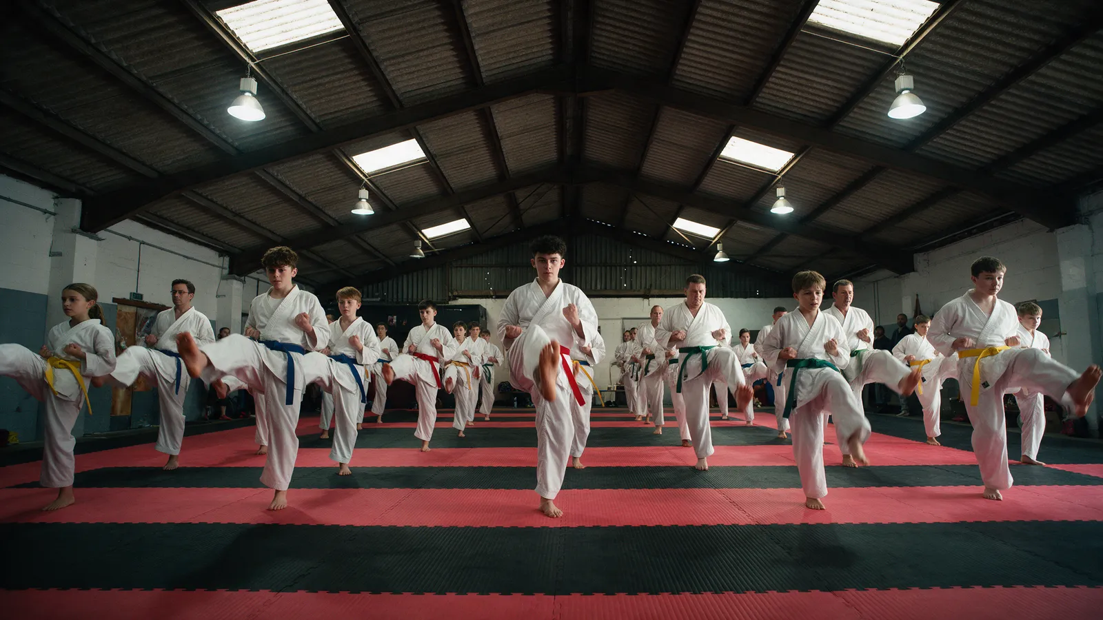 A karate class of teenagers and adults in white gis with coloured belts performing a synchronised front kick in rows on red and black mats