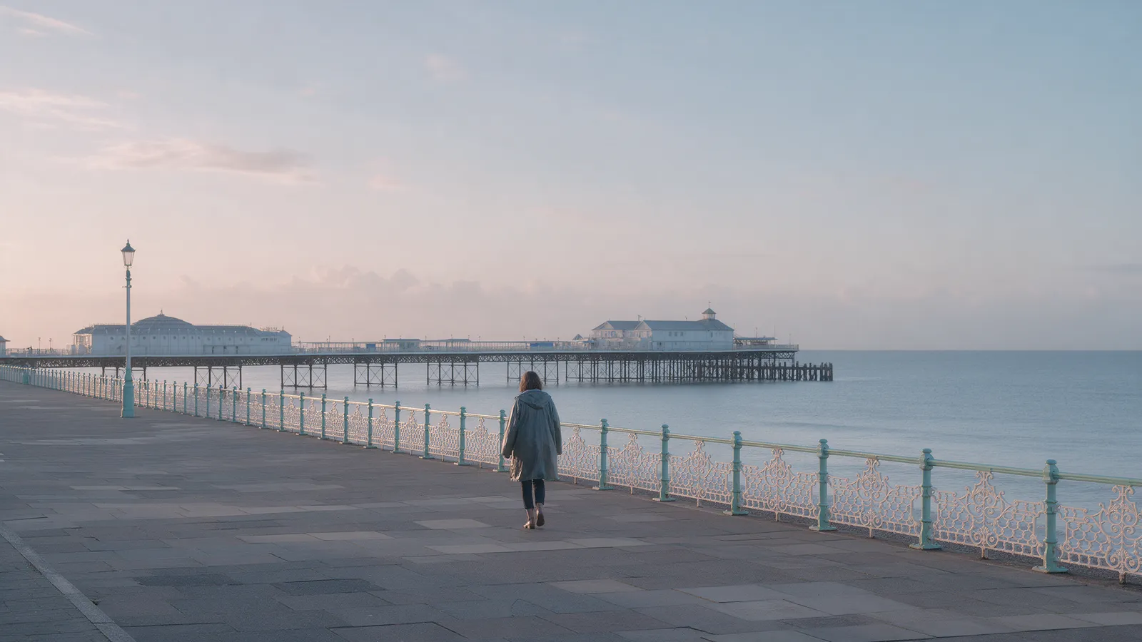 A woman in a raincoat walking alone along a Victorian seaside esplanade at dawn with a long pier and a calm grey-blue sea
