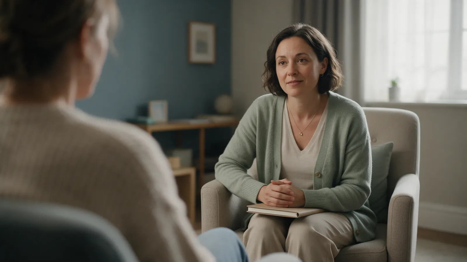 A counsellor in a sage cardigan listening in an armchair, seen over the shoulder of a client in a calm room with soft window light