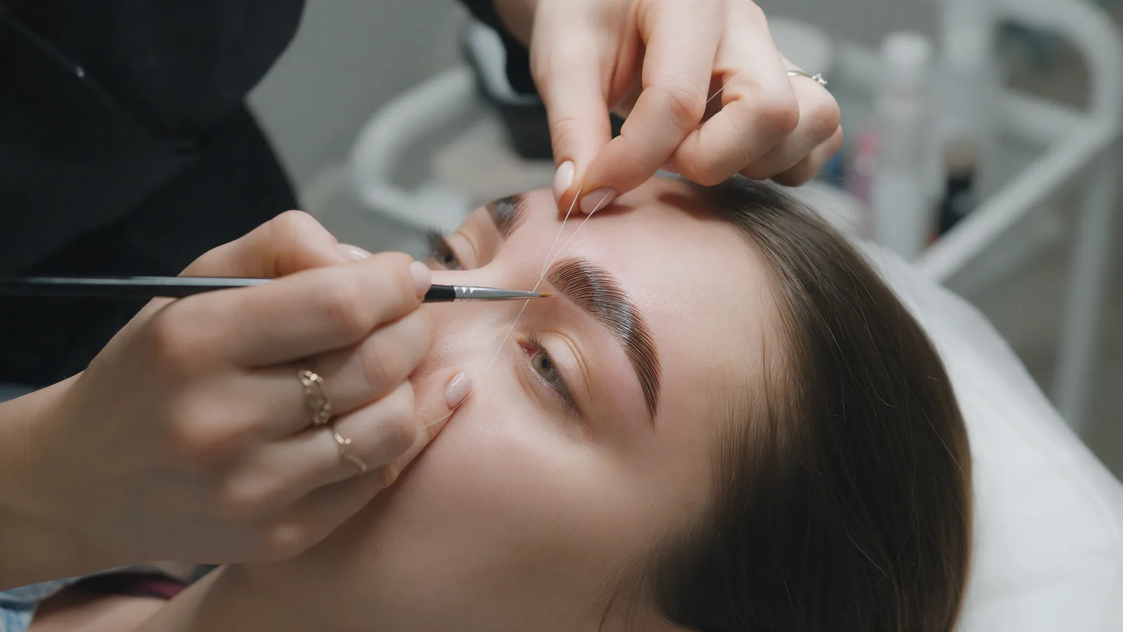 A brow technician mapping and shaping a client's eyebrows with a fine brush, the brows laminated and defined