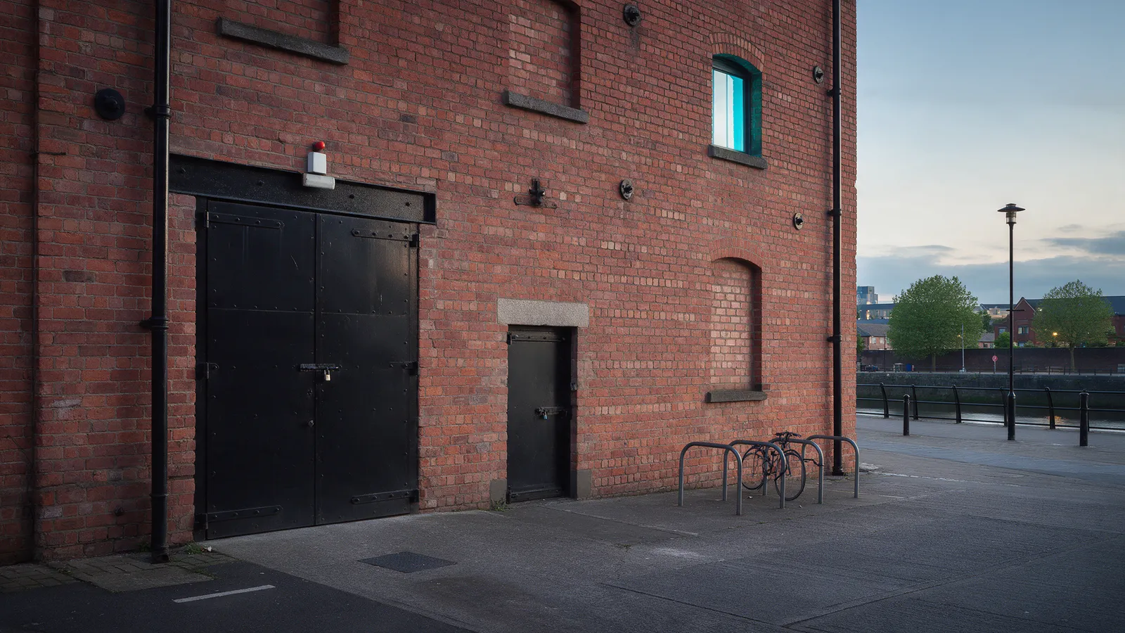 The exterior of a converted red-brick Victorian warehouse unit beside a quiet river embankment, a heavy black steel door and a loading door, a bicycle rack, evening light