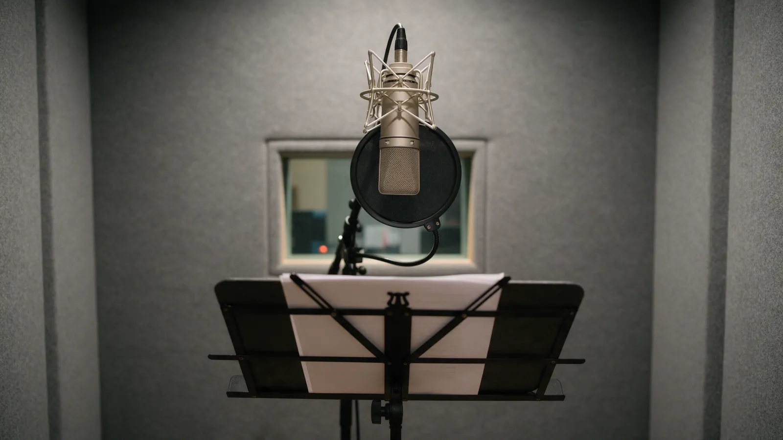 A voice-over booth with a large-diaphragm condenser microphone, a pop shield, a music stand and grey acoustic foam walls, a small window to the control room