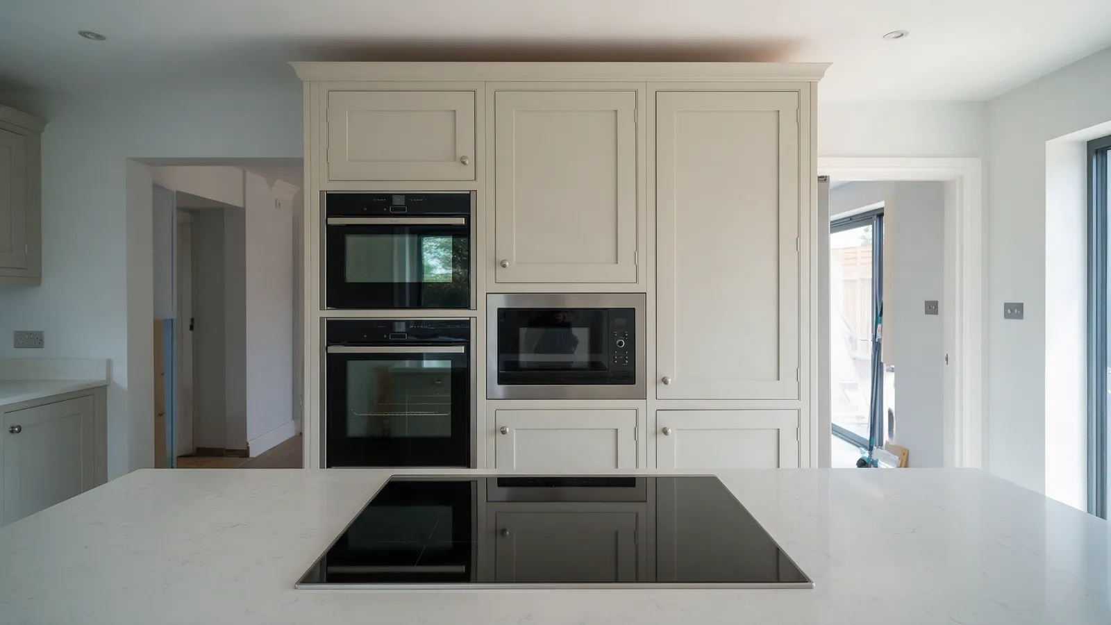 Two stacked built-in ovens and a combination microwave in pale linen tall cabinets beside an integrated fridge, an induction hob on a quartz worktop in front