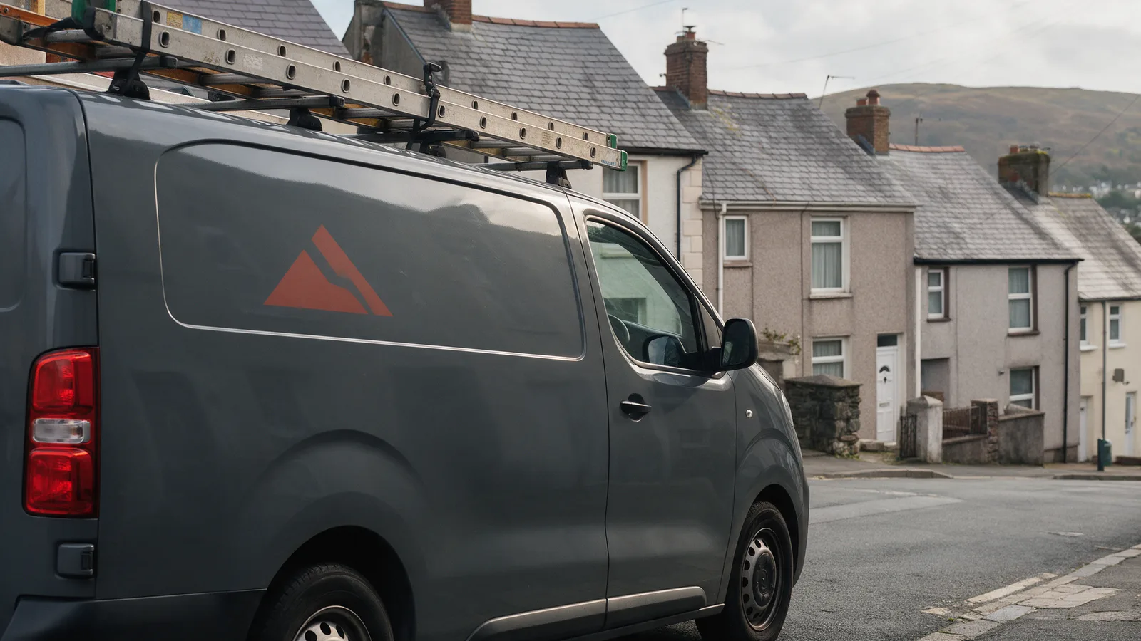 A dark grey Ridgeline Roofing van with ladders on the roof rack parked on a Welsh terraced street