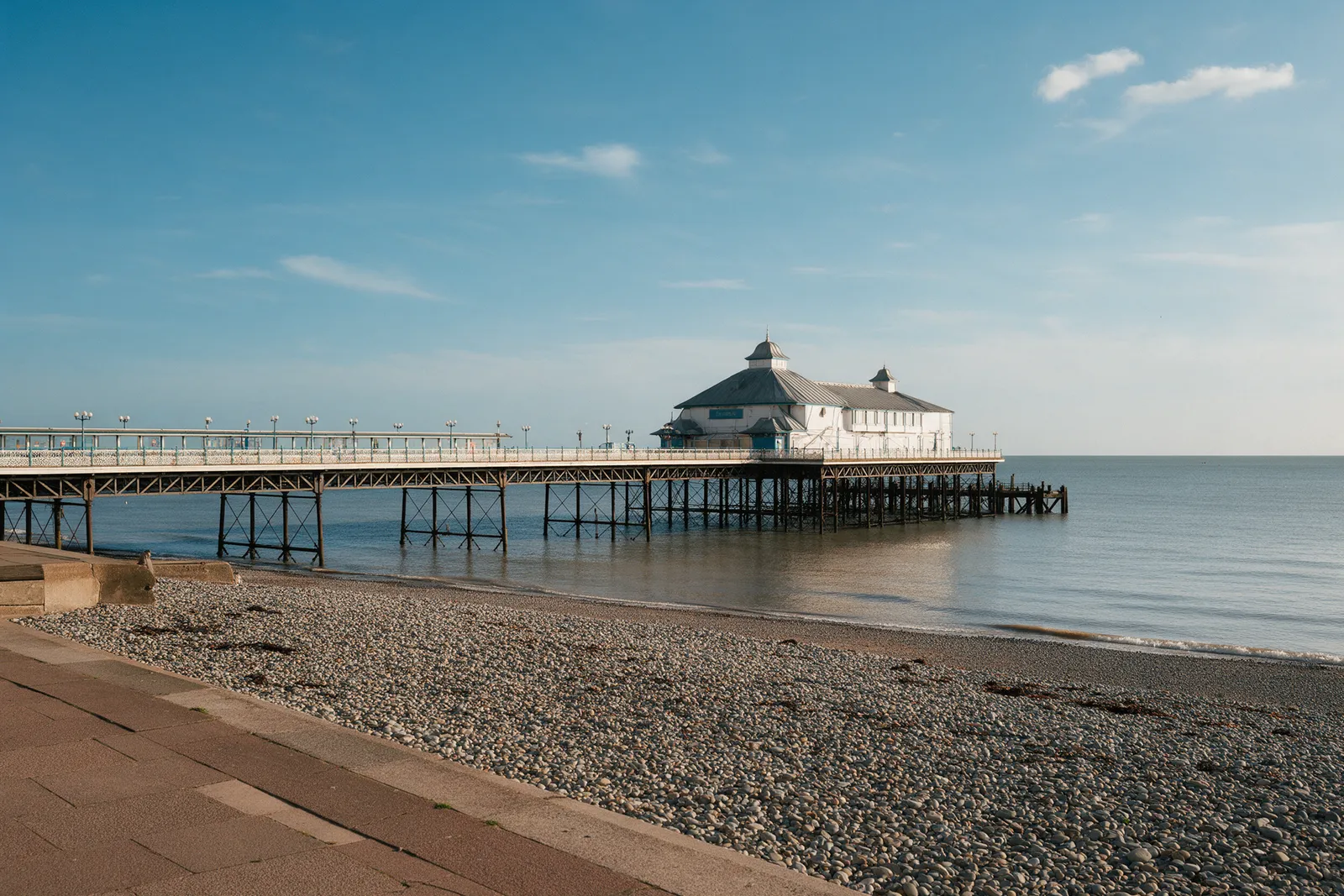 A Victorian seaside pier with a pavilion stretching into a calm sea, a pebble beach and esplanade in the foreground