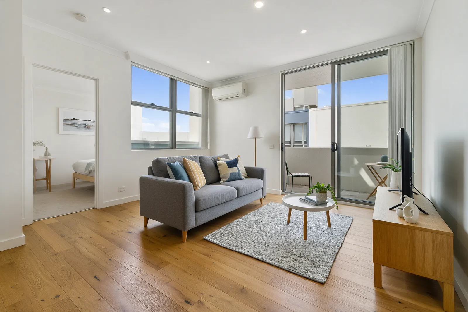A bright modern two-bedroom flat living room with a grey sofa, oak floor, large windows and a small balcony