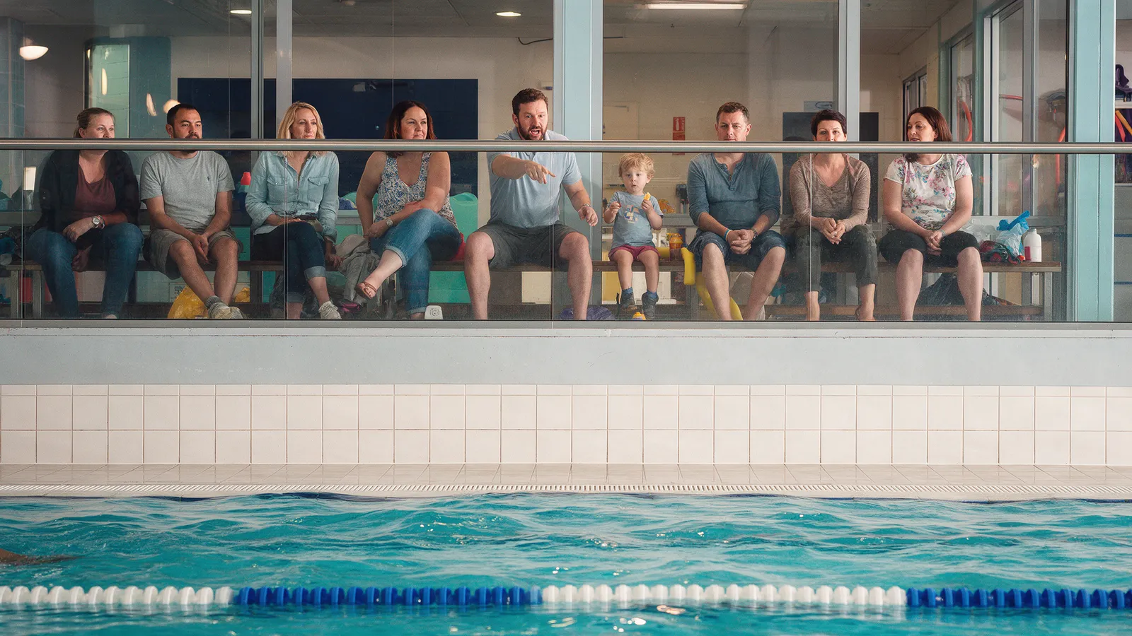 Parents on benches behind a glass balcony watching swimming lessons in the pool below