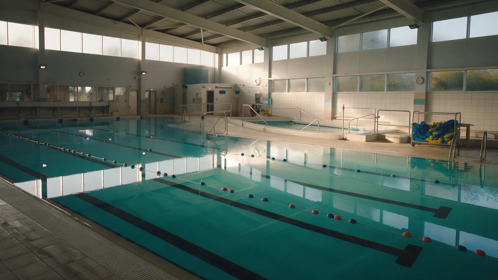 An empty indoor 25-metre pool early in the morning with lane ropes laid out and a separate teaching pool with steps at the side