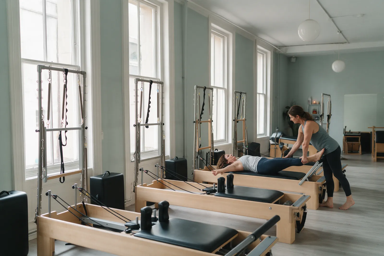 A Pilates reformer studio with four reformer machines in a row by tall windows, a woman lying on one with a teacher adjusting her foot position
