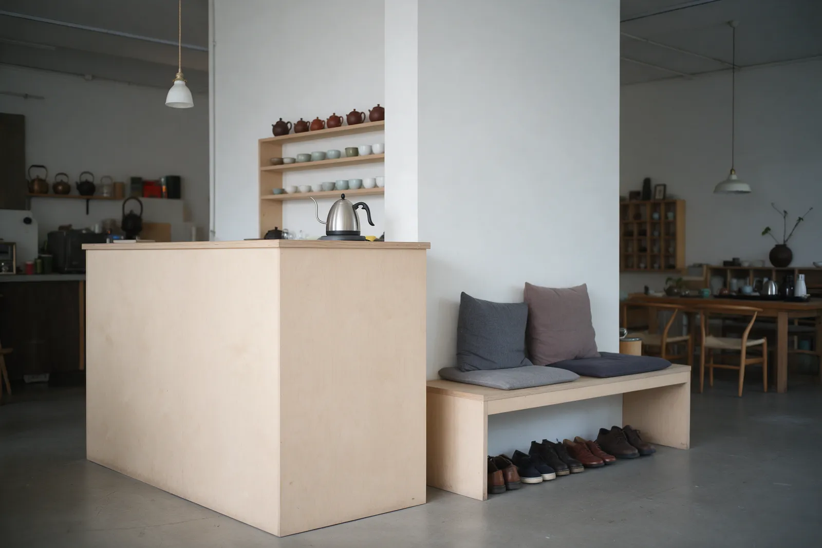 A quiet studio reception and tea corner with a pale wooden counter, a kettle, a shelf of teapots and cups, a bench with cushions and shoes lined up neatly beneath