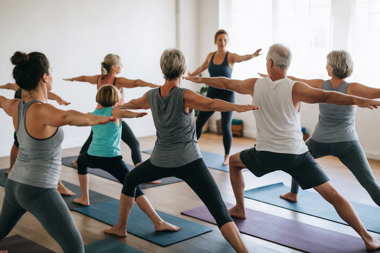 A small yoga class of six people of mixed ages in warrior pose on mats in a bright studio with a teacher at the front