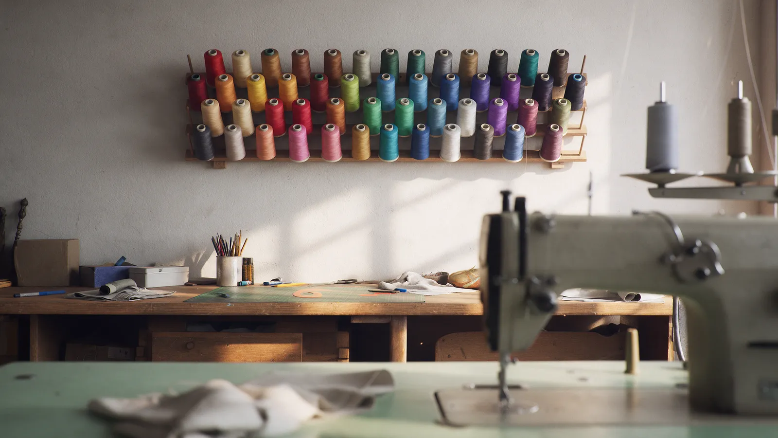 A wall rack of thread spools in every colour above a workroom bench, an industrial sewing machine in the foreground