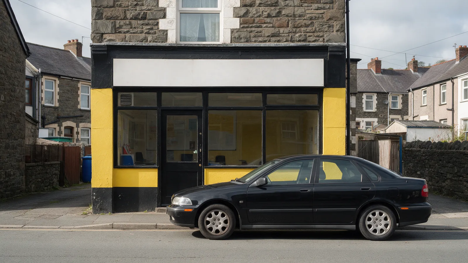 A small taxi office with a black and yellow painted frontage on a stone-built Valleys street, a saloon car parked outside, no readable text