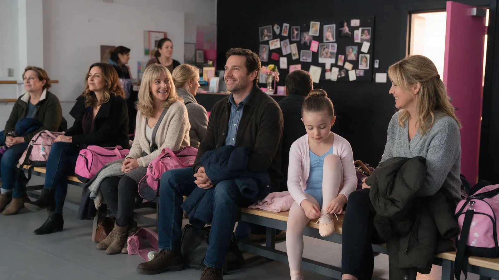 Parents on a bench in a dance school reception with dance bags, a small girl tying her ballet shoe