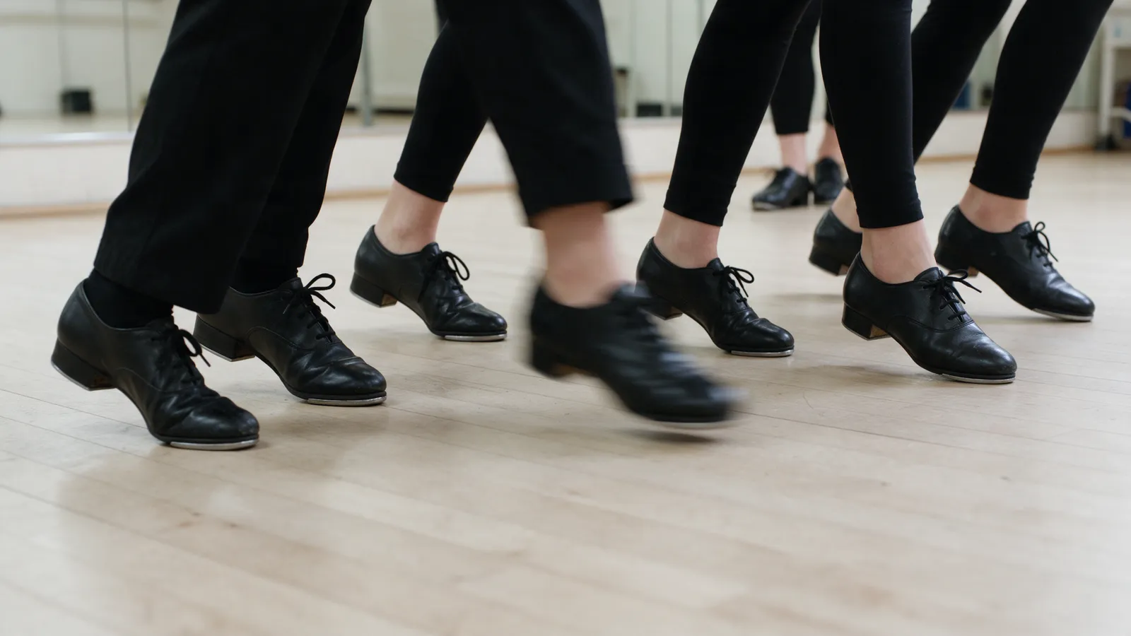 Close-up at floor level of six pairs of black tap shoes mid-step on a pale sprung wooden floor, a mirrored wall behind