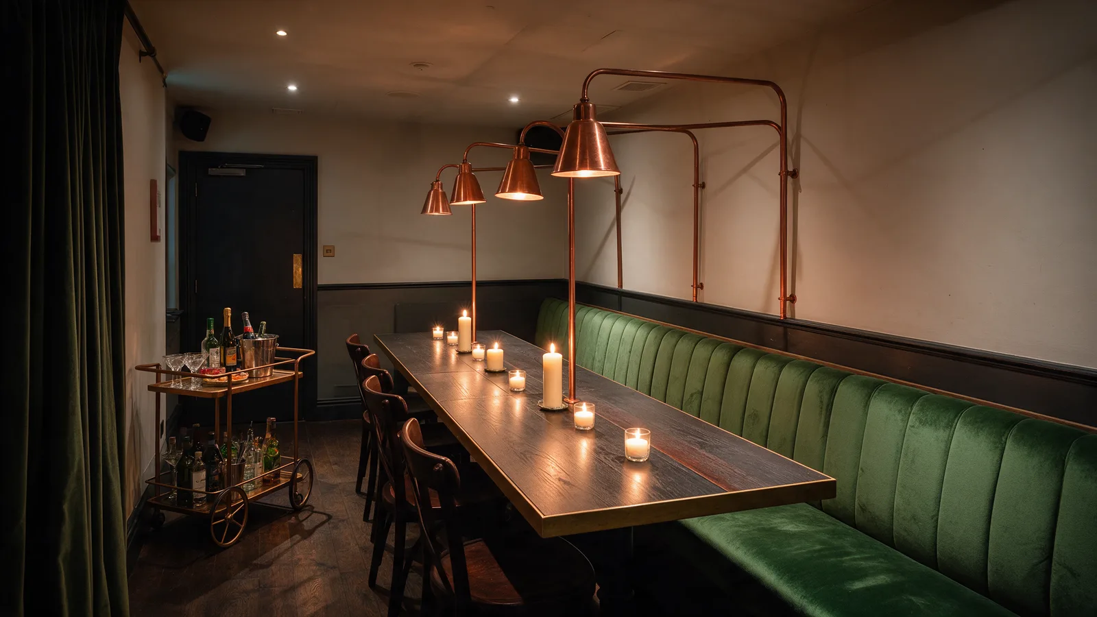 The back room set for a party: a long table with candles, a green velvet banquette, copper lamps and a drinks trolley