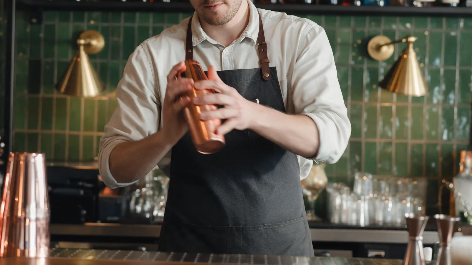 A bartender in a dark apron shaking a cocktail in a copper shaker behind a bar with a green tiled wall