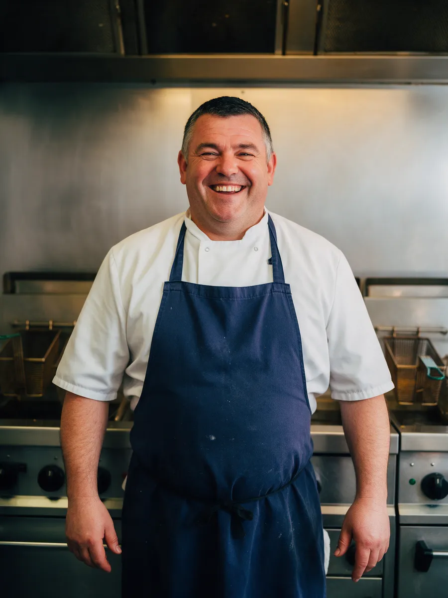 Dai Price, a cheerful Welsh man in his late forties with short dark hair greying at the sides, in a white tunic and navy apron in front of a stainless steel frying range