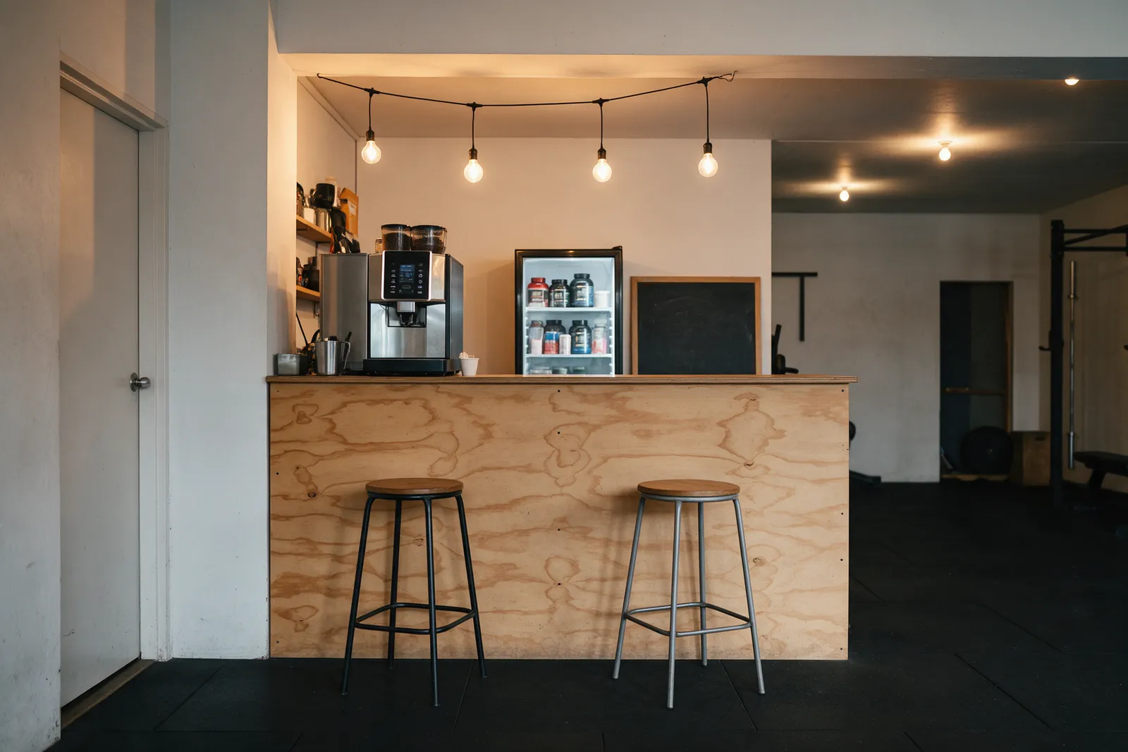 A gym reception with a plywood desk, a coffee machine, a protein-shake fridge, two stools and a chalkboard, warm welcoming light