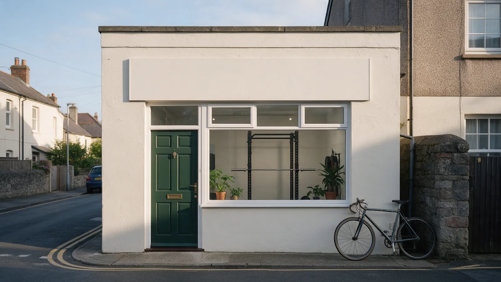 The exterior of a small private training studio in a converted mews on a quiet Penarth side street: a dark green door, a large window showing a squat rack and plants, a bicycle